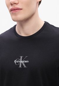 T-shirt preta de algodão com um logótipo bordado cinza com "Calvin Klein" e "CK" na parte da frente, decote redondo, textura suave.