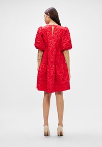 Raudona su pūstomis rankovėmis suknelė su tekstūruotu paviršiumi, sluoksniuotu volanų sijonu ir atviru nugara. Derinama su nuoga aukštakulnių batais.