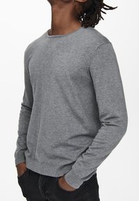 Homme portant un pull gris uni à manches longues avec les mains dans les poches d'un jean noir, sur un fond blanc uni.