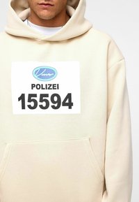 Beige hoodie met een frontzak, groot wit label met "POLIZEI 15594" in het zwart, en een blauw logo boven het label.
