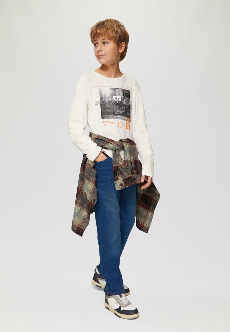 Ciemnobeżowa koszulka z długim rękawem z graficznym nadrukiem, niebieskie dżinsy i wozownica w kratę związana w talii. Białe i czarne sneakersy.