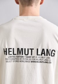T-shirt en coton beige clair avec un col rond. Le dos présente un logo en texte noir, incluant "HELMUT LANG" et des détails supplémentaires dans une police audacieuse.
