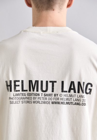 Kerges beež puuvillane T-särk ümarate kaelustega. Seljal on musta teksti brändimine, sealhulgas "HELMUT LANG" ja täiendavad detailid rasvases fondis.