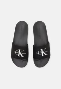 Svarte sandaler med skliddesign, som har en myk gummisåle og en stoffrem prydet med en hvit "CK"-logo og teksten "Calvin Klein Jeans".