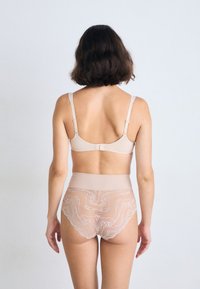 Γυναίκα με καστανά μαλλιά μέχρι τους ώμους φορώντας μπεζ σουτιέν και κορσέ με δαντέλα ψηλά στη μέση, όρθια ενάντια σε μια απλή ανοιχτόχρωμη επιφάνεια, με την πλάτη στραμμένη προς την κάμερα.