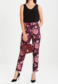 Zwarte mouwloze top gecombineerd met bloemenbroek met elastische taille in roze en paarse tinten. Model houdt een bordeauxrode handtas vast. Zwarte hakken.