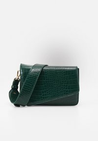 Mörkgrön texturerad handväska med en lång rem, med en elegant design och glänsande yta, uppställd mot en ljus bakgrund.