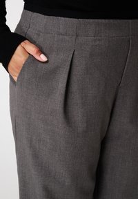 De hand van een vrouw met lichtroze nagellak in de grijze geplooide zak van een op maat gemaakte broek, en een zwarte geribbelde mouw zichtbaar boven de tailleband.