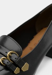 Μαύρο δερμάτινο λουστρίνι με γυαλιστερή φινίρισμα, διαθέτει μεταλλικά διπλά κούμπωμα σε χρυσό τόνο και μυτερή μύτη. Το εσωτερικό έχει καφέ επένδυση.