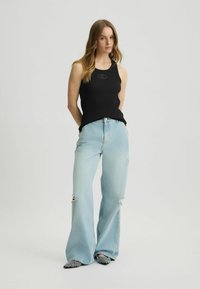 Top de tirantes negro con diseño acanalado que presenta un logo en el pecho; combinado con unos vaqueros de pierna ancha de color azul claro con dobladillo deshilachado y detalles desgastados.