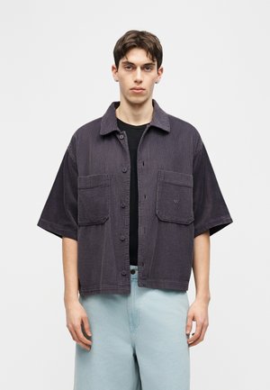 Jonge man die een donkergrijs kort mouw, corduroy overhemd met knoopjes draagt over een zwart shirt en lichtblauwe spijkerbroek, staand tegen een effen achtergrond.