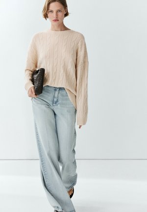 Kvinde iført en beige kabelstrikket sweater, lyseblå brede jeans, sorte sandaler og holder en sort flettet clutch.