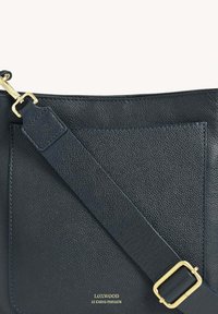 Sac à main en cuir bleu marine avec une surface texturée, une bandoulière réglable et des accessoires en métal doré, comprenant une poche avant et un détail de logo.