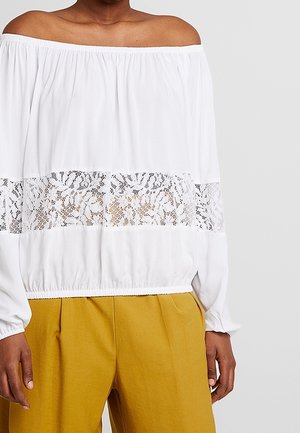Biała bluzka z odkrytymi ramionami i koronkowym panelem na wysokości piersi, zestawiona z musztardowo-żółtymi, wysokim stanem spodniami na osobie.