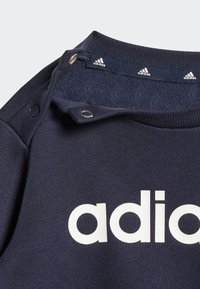 Felpa navy con una texture morbida, decorata con il logo "adidas" bianco. Chiusura a scatto sul colletto con tre piccoli accenti del logo sul rivestimento.