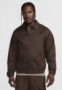 Brun zip-up jakke med krave, elastiske manchetter og et diskret Nike-logo. Har en glat, mat finish og et afslappet fit design.