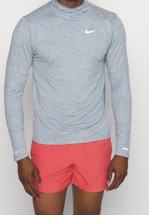 Mężczyzna ubrany w dopasowaną jasnoszarą koszulkę Nike Dri-FIT z długim rękawem i czerwone krótkie spodenki sportowe, stojący na jednolitym tle.