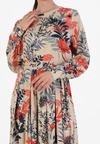 Geblümtes Kleid mit langen Ärmeln, beiger Stoff mit roten, dunkelblauen und grünen Blumen; plissierter Rock und passender Gürtel in der Taille.