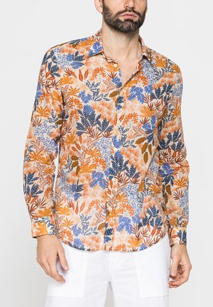 Uomo che indossa una camicia a maniche lunghe con bottoni, con un motivo floreale colorato arancione, blu e marrone e pantaloni bianchi.