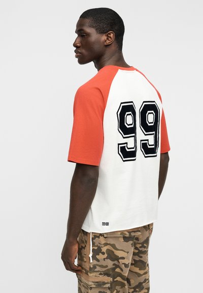 Κοντομάνικο T-shirt κόκκινου και άσπρου χρώματος με μεγάλο μαύρο «99» στην πλάτη. Συνδυάζεται με χακί cargo σορτς με πολλαπλές τσέπες.