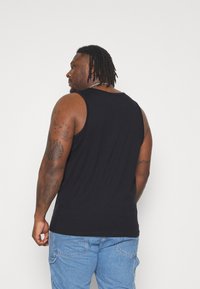 Top nero senza maniche, realizzato in un tessuto morbido, dal taglio aderente, con tatuaggi visibili sul braccio. Indossato con pantaloni in denim chiaro.