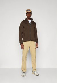 Casaco de flanela castanho com fecho éclair, calças castanhas claras e sapatilhas bege. Camisola interior branca e boné bege com logótipo. Tecido liso, ajuste descontraído, sem padrões.