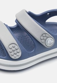 Modré sandály s texturovaným designem, dvěma šedými nastaviteľnými popruhy s vyvýšenými tečkami a bílou podrážkou s detailním logem.