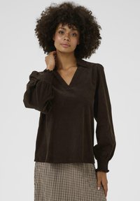 Blouse en velours côtelé marron avec un décolleté en V, des manches bouffantes et des poignets smockés. Présente une texture lisse et une coupe décontractée.