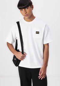 White cotton t-shirt with short sleeves and a chest pocket bearing a black label. Model holds a small black crossbody bag.