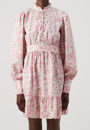 Kvinde iført en hvid kjole med lange ærmer og høj hals, dekoreret med pink blomsterprint, knappet foran, tilpasset talje og med flæsekant over knæene.