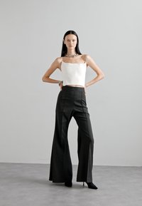 Fehér textúrájú crop top, négyzet formájú nyakkivágással, fekete csíkos bővülő nadrággal párosítva. A modell egy szürke háttér előtt áll.