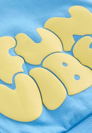 Sininen puuvillakangas, jossa on suuria keltaisia kumimaisia kirjaimia pyöristettyine reunoineen ja tyyliteltyä muotoilua muistuttavia yksityiskohtia.