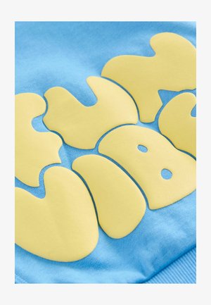 Modra bombažna tkanina z velikimi rumenimi gumo obdelanimi črkami zaobljenih robov in detajli, ki spominjajo na stilizirano zasnovo.