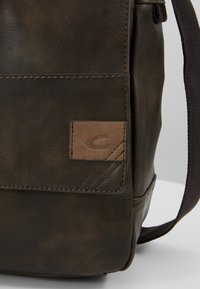 Dunkelbraune Leder-Schultertasche mit strukturiertem Finish, ausgestattet mit einem kleinen, hellbraunen Lederpatch und Nahtdetails. Verstellbarer Stoffgurt inklusive.