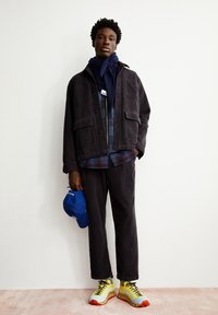 Zwarte corduroy jas met opgestikte zakken, gedragen over een geruite blouse. Bijpassende broek, blauwe pet en gele sneakers met rode accenten.