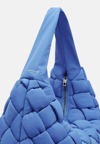 Niebieska torba tote z pikowanym wykończeniem, z dwiema rączkami i zapięciem na zamek błyskawiczny. Nazwa marki "LIEBESKIND" jest widoczna na torbie.