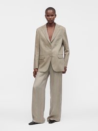 Mannequin mince à la peau foncée portant un costume beige à fines rayures oversize avec pantalon à jambes larges et chaussures noires pointues, sur un fond uni.