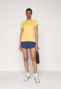 Νεαρή γυναίκα με κίτρινη αθλητική μπλούζα, μπλε σόρτς και λευκά αθλητικά παπούτσια κρατώντας μαύρη μπουκάλι νερού, σε απλό φόντο.