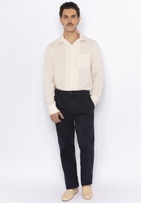 Chemise à manches longues crème avec poche, associée à un pantalon sombre et des mocassins clairs. Texture lisse, coupe classique, design de col classique.