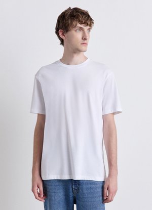 Giovane uomo con capelli castano chiaro che indossa una semplice maglietta bianca a maniche corte e jeans blu su uno sfondo chiaro uniforme.