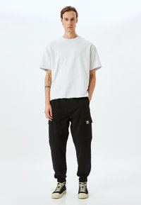 Witte oversized t-shirt met rauwe randen, gecombineerd met zwarte cargobroek met zijzakken en elastische boorden. Zwarte hoge sneakers met een crèmekleurige zool.
