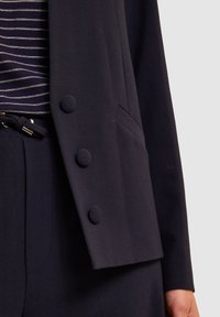 Granatowa marynarka z trzema okrągłymi, pokrytymi tkaniną guzikami, jedną kieszenią na piersi i miękką teksturą. Nosi się ją na paskowanym topie.