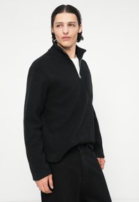 Pulover negru din fleece cu guler cu fermoar pe o pătrime și material texturat. Prezintă o buzunar frontal și mâneci lungi. Asortat cu pantaloni negri.