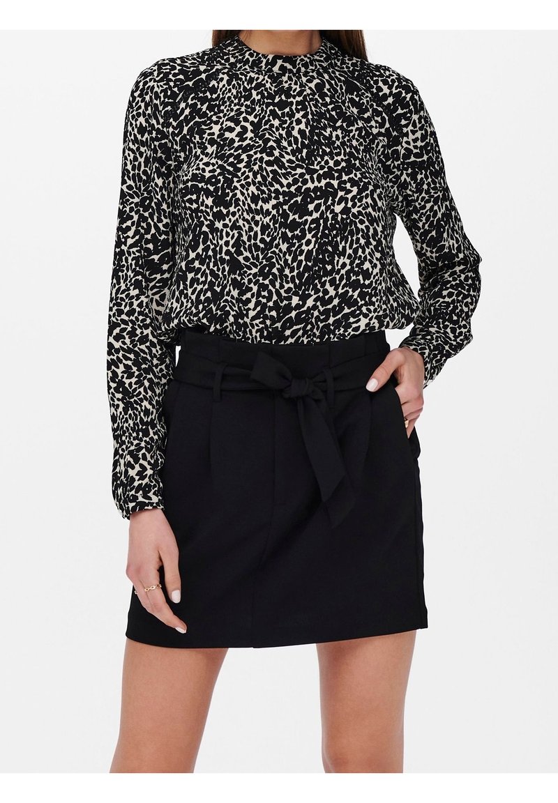 Femme portant un chemisier à manches longues imprimé animal noir et blanc, rentré dans une jupe noire taille haute avec une ceinture nouée.