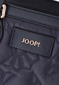 Tummansininen tilkkukangas, jossa on kohotettuja ommelkuvioita. Varustettu suorakulmaisella mustalla tarralla, jossa on kohokohtaisena kultainen "JOOP!"-logo.