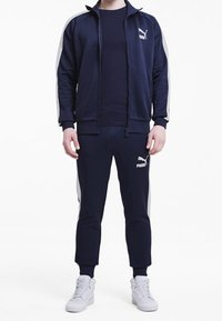 Námornický teplákový komplet vyrobený z hladkého materiálu, obsahující zapínací bundu s bílými pruhy po stranách a odpovídající kalhoty. Logo viditelné na obou kusech.