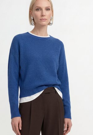 Kvinde iført en blå strikket sweater over en hvid skjorte, kombineret med mørkebrune bukser og store runde sølvøreringe, står foran en hvid baggrund.