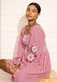 Vrouw zittend in een rood-wit geruite jurk met bloemenborduursels op de mouwen, voor een lichte bakstenen muur.