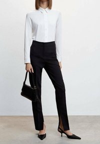 Weißes Langarmshirt mit Knopfverschluss, kombiniert mit hoch taillierten schwarzen Hosen mit Seitenschlitzen und spitzen schwarzen Absätzen, ergänzt durch eine kleine schwarze Handtasche.