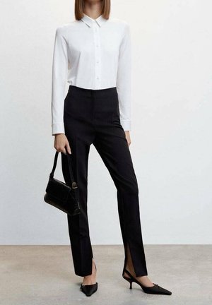 Weißes Langarmshirt mit Knopfverschluss, kombiniert mit hoch taillierten schwarzen Hosen mit Seitenschlitzen und spitzen schwarzen Absätzen, ergänzt durch eine kleine schwarze Handtasche.